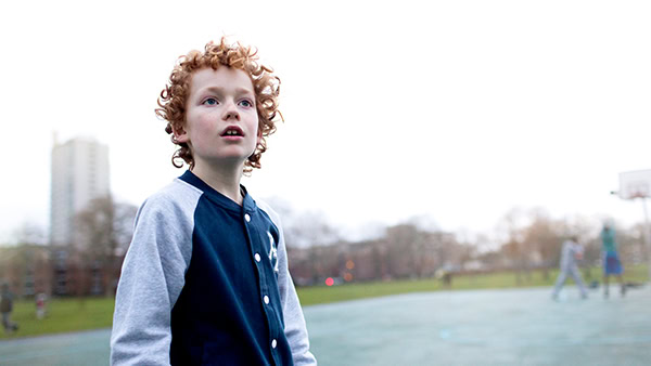 A young boy with curly red hair, wearing a blue and gray jacket, stands on an outdoor basketball court. The background shows a tall building and blurred figures playing basketball in the distance. The sky is overcast.