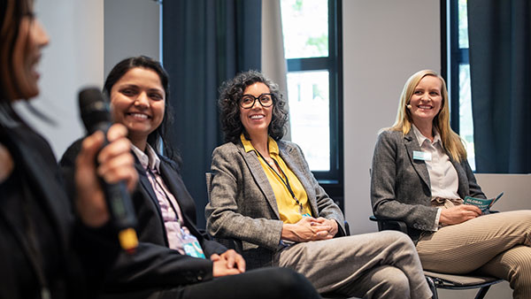 Four women in business attire sit and engage in a panel discussion. One holds a microphone while the others smile and listen. They are in a modern conference room with large windows and curtains.