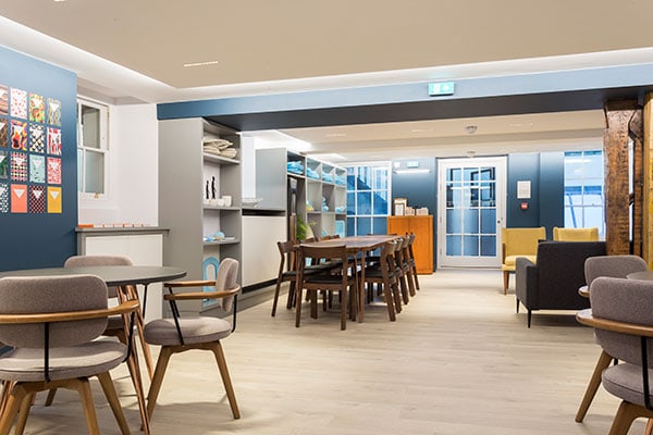 A modern, well-lit interior with gray and wood-toned furniture. A long wooden dining table with chairs is centered. Seating areas with armchairs and circular tables are on either side. Art decorates blue walls, and large windows provide natural light.