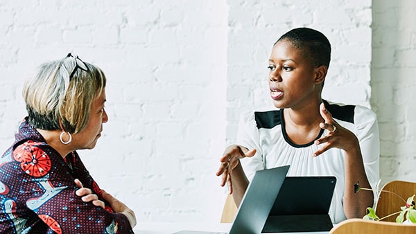 Two women sit at a table in conversation. One woman gestures with her hands while speaking, and an open laptop is on the table. The setting is bright with a white brick wall in the background.