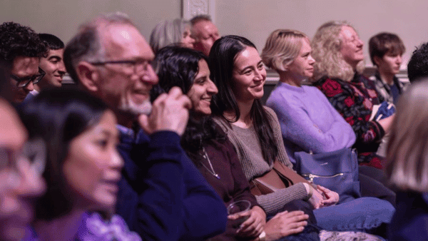 A diverse group of people sitting in an audience, smiling and listening attentively. The setting appears to be a seminar or presentation. Everyone is dressed casually, and the atmosphere seems engaging and positive.