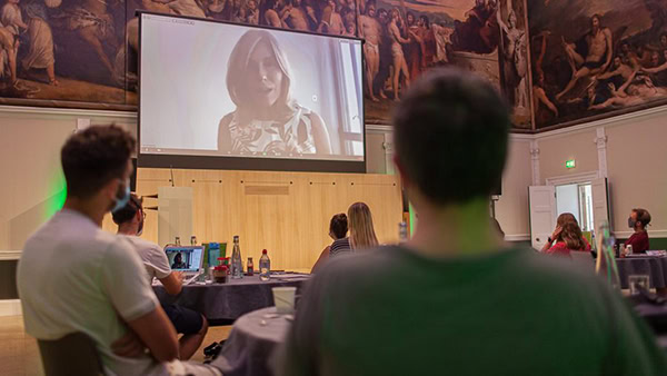 People seated in a room with ornate wall paintings, watching a woman speaking in a large projected video at the front. The room has dim lighting, and some attendees have laptops.