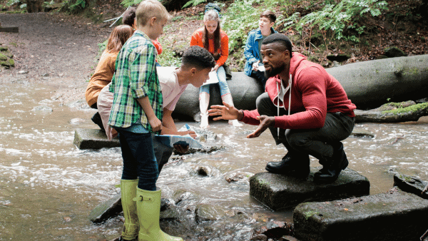 A group of children and a man are outdoors by a creek. The man is crouching on a rock, speaking to a boy in a plaid shirt and green boots. Other children are sitting on rocks and logs, observing. The setting is a wooded area.