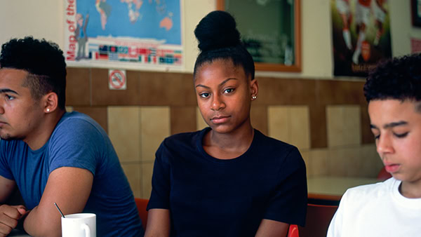 Three people sit at a table in a cafe. The person in the center looks forward, wearing a black top and hair in a bun. Another wears a blue shirt, and the third wears a white shirt. There's a world map on the wall behind them.