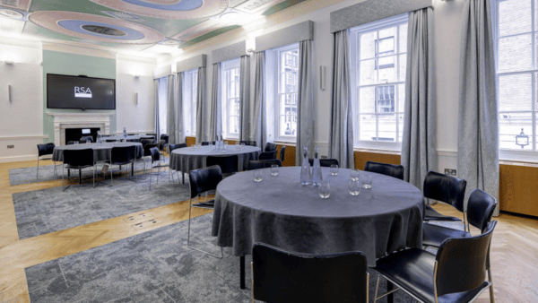 Elegant meeting room with round tables covered in gray tablecloths, surrounded by black chairs. Tall windows with gray curtains allow natural light. A large screen is mounted on a pastel-colored wall, featuring a modern design and wood flooring.