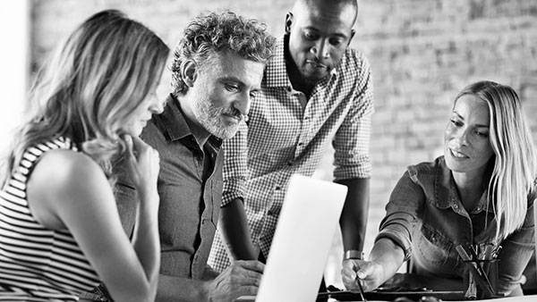 Four people are gathered around a laptop, intently discussing something. Two are seated, while two stand. They are focusing on the screen, with one person gesturing toward it. The setting has a casual, collaborative atmosphere.