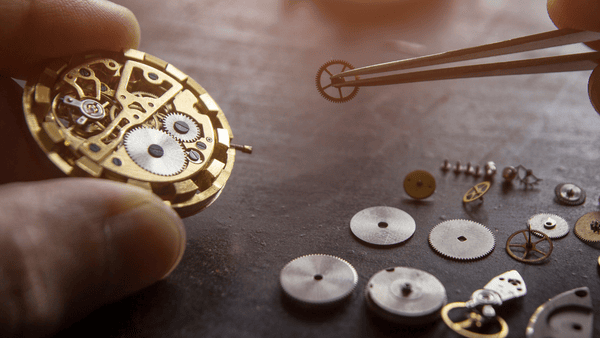 Close-up of a person's hands assembling a mechanical watch. One hand holds the watch mechanism, while the other uses tweezers to place a small gear. Various gears and parts are scattered on a dark surface.
