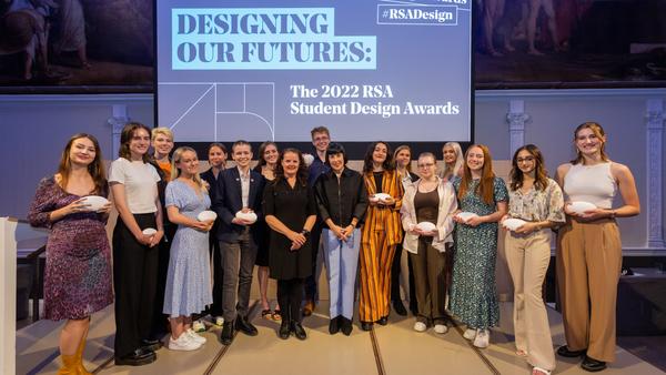 A group of people posing on a stage in front of a screen displaying "Designing Our Futures: The 2022 RSA Student Design Awards." Some individuals are holding white objects. The background features large paintings.