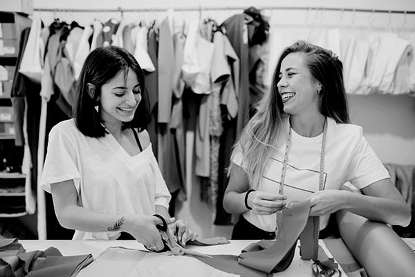 Two smiling women work at a table with fabric and scissors in a sewing studio. One woman holds a piece of cloth while the other cuts it. Shelves with hanging garments are visible in the background. Black and white image.