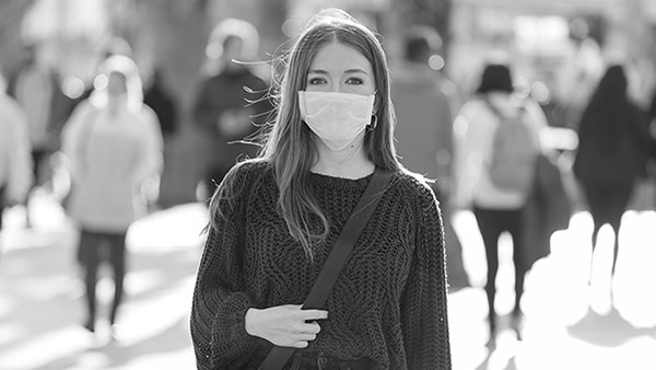 A woman wearing a face mask stands outdoors with a shoulder bag. She's surrounded by a blurred crowd in the background. The setting appears to be a sunny urban street. The image is in black and white.
