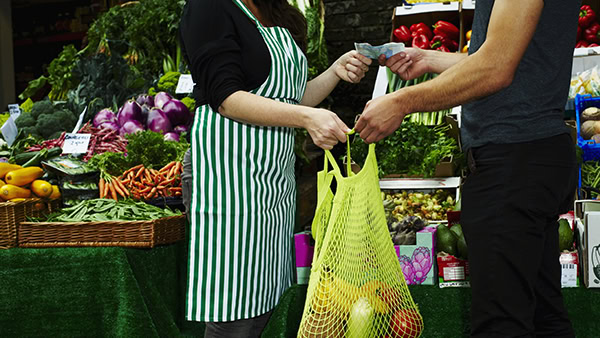 A vendor in a striped apron hands a mesh bag filled with vegetables to a customer at an outdoor market stall. The stall is stocked with various fresh produce, including carrots, zucchini, and peppers. The customer is paying with cash.