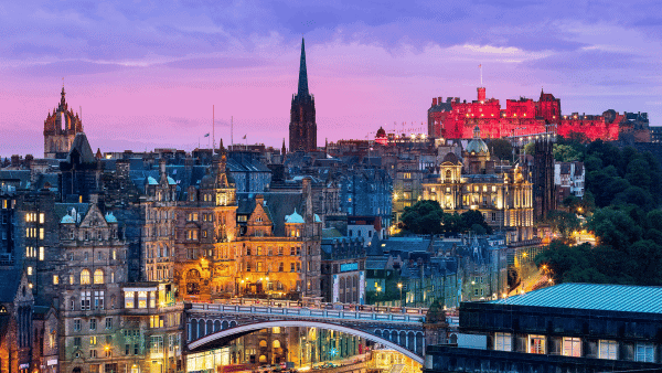 A scenic view of Edinburgh at dusk, featuring historic buildings with warm lights. The prominent Edinburgh Castle is illuminated atop a hill, while church spires punctuate the skyline under a pink and purple sky.