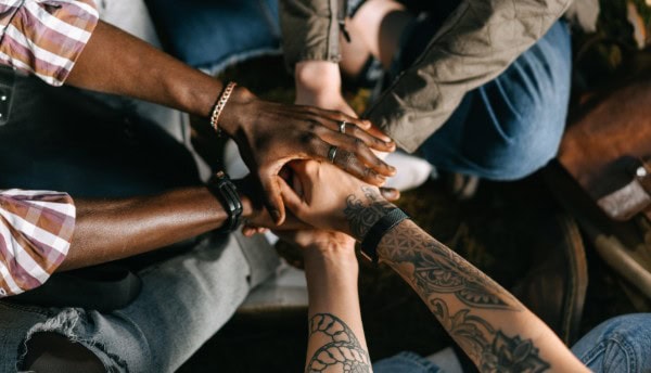 A group of people sitting in a circle places their hands together in the center, symbolizing social connection. Their hands vary in skin tone and have different accessories and tattoos, representing unity and diversity.