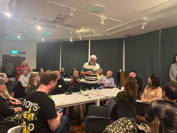 A group of people sit and stand around tables in a well-lit school room, engaged in discussion. One man, standing in the center, appears to be addressing gender stereotyping. Papers and drinks are on the tables.