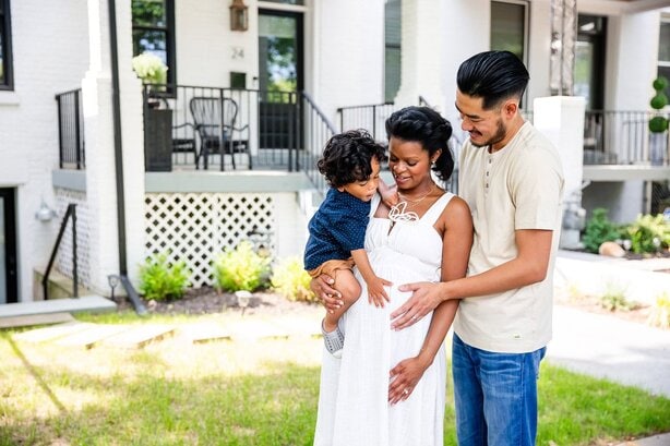 During an on-location Washington DC family photo shoot, a pregnant mother stands on the sidewalk while holding her toddler son. She is being embraced from behind by her husband while he and toddler son touch the her belly.