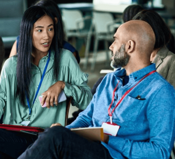 A woman and a man wearing lanyards sit and talk at the Royal Society for the Arts. The woman holds a phone and gestures as she speaks, while the man listens attentively. Other people are blurred in the background.