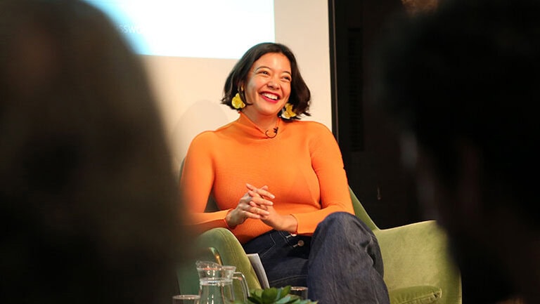 A person wearing an orange sweater and sitting in a chair smiles while engaging in a conversation. They have short hair and are wearing yellow earrings. The room appears to be a setting for a talk or presentation, with a blurred audience in the foreground.