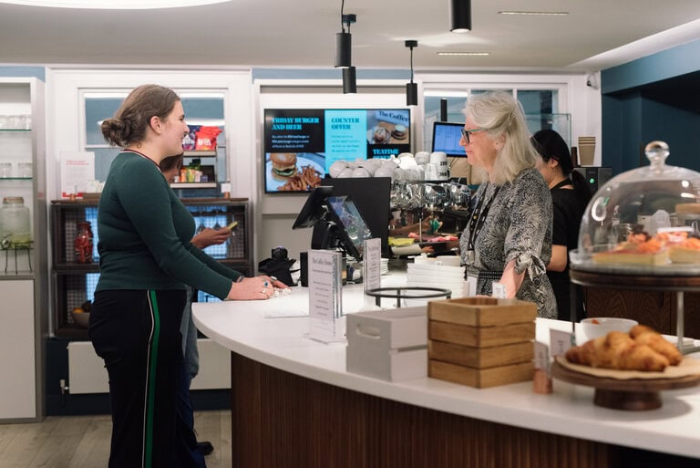 A woman with glasses stands behind a modern café counter, discussing ideas with another woman in a green sweater. Nearby are pastries under a glass dome and digital screens displaying menu items, creating an atmosphere reminiscent of the innovative spirit found at the Royal Society for the Arts.