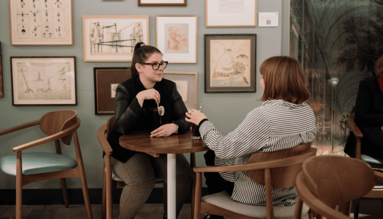 Two women are seated at a small round table in a cozy cafe. They appear to be engaged in conversation, with one holding a cup. The wall behind them is decorated with various framed artworks.