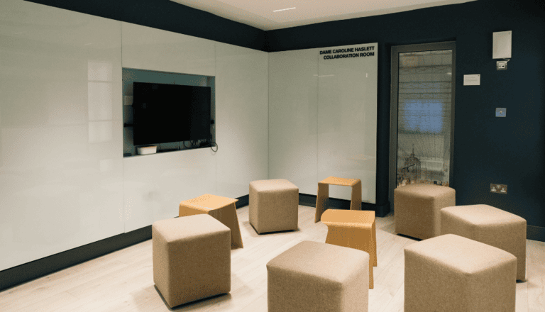A modern meeting room with a large screen mounted on a whiteboard wall. Beige cube and hexagonal stools are arranged randomly on a light wood floor. A door with a glass panel is on the side. The room is lit with recessed lighting.