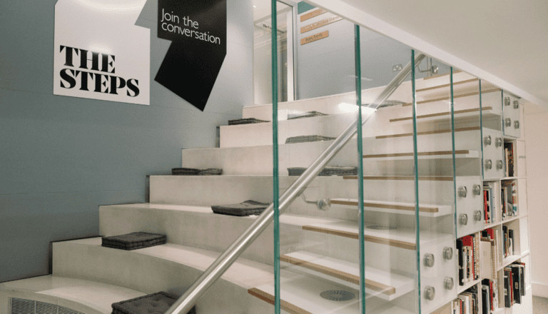 Indoor staircase with glass side panels and wooden steps adorned with cushions. A wall sign reads "THE STEPS" and "Join the conversation." Bookshelves filled with books are visible along the staircase.