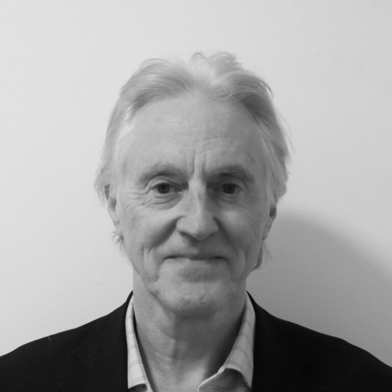 Black and white photo of an older man with light hair, wearing a dark blazer over a light, striped shirt. He is smiling slightly and is standing against a plain, light-colored background.