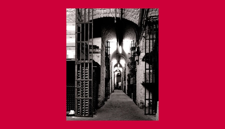A black and white photo of a narrow, dimly lit corridor with stone walls and archways. Steel gates with vertical bars line both sides, resembling prison cells. The scene is framed by a red border.