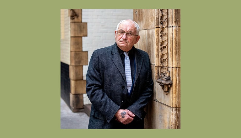 Elderly man in a dark suit and tie, leaning against an ornate, carved stone pillar. He has short white hair, glasses, and is looking to the side. The background has white and black tiled walls, and the image is framed with a green border.