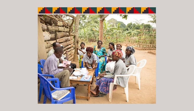 A group of people sit outdoors in a circle on plastic chairs, engaged in discussion. Papers and notebooks are on a central table. A wooden wall and leafy background are visible. The top border features a pattern of red and black geometric shapes.