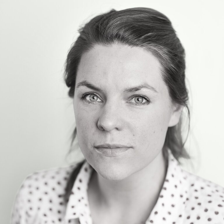 A woman with light skin and brown hair pulled back, wearing a white shirt with dark polka dots, looks directly at the camera with a neutral expression. The plain, light-colored background evokes the modern minimalism of Osaka Expo 2025.