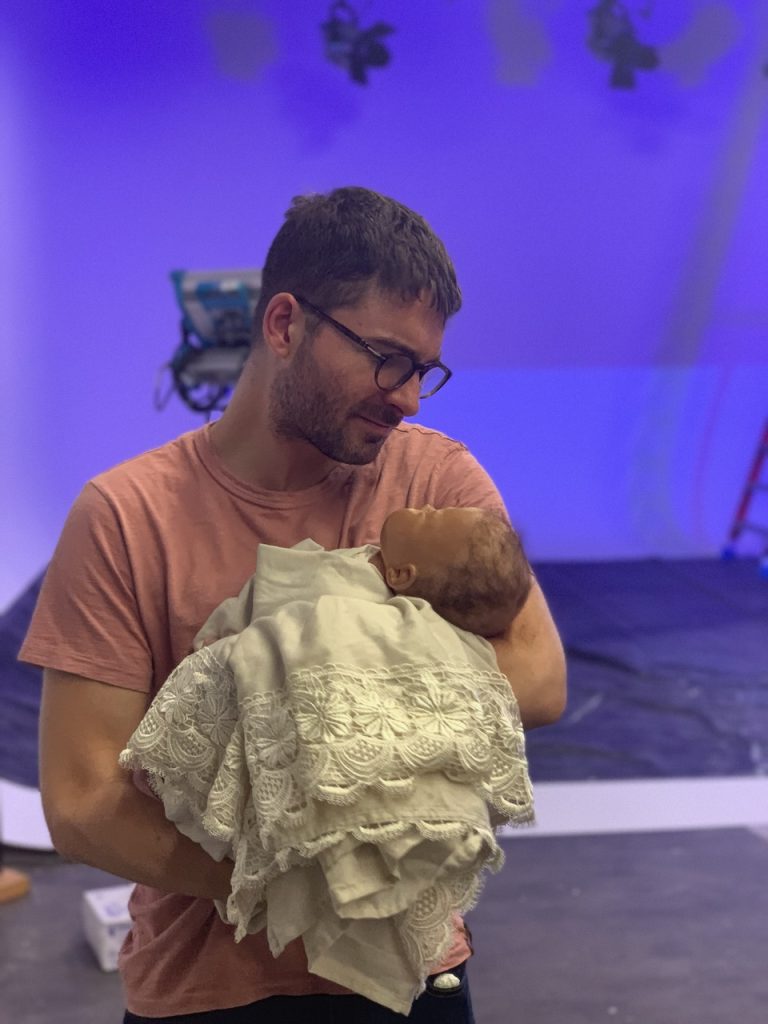 Director in peach shirt and glasses cradles swaddled infant on Tulsa Opera commercial set with purple lighting