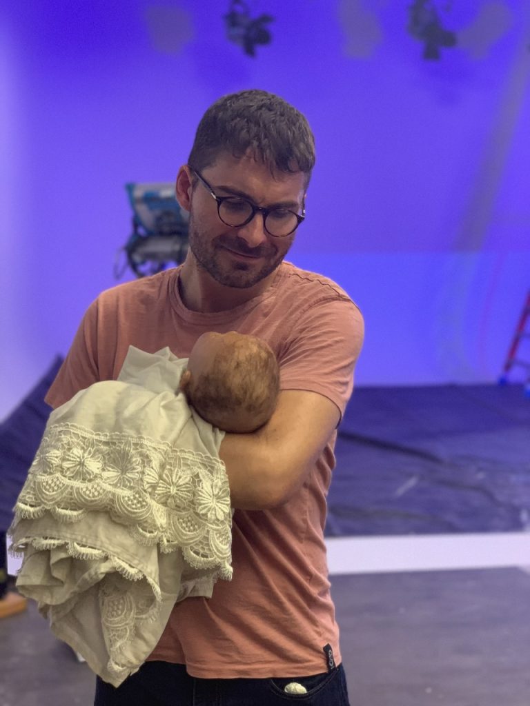 Director in glasses and orange shirt holds white lace-wrapped doll on Tulsa Opera campaign set with blue stage lighting