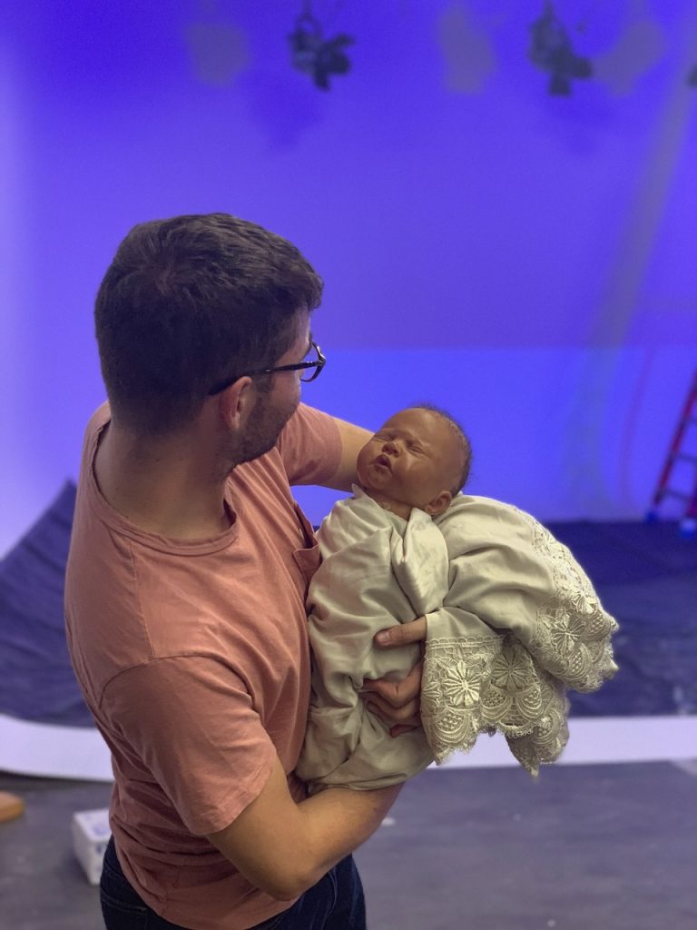Man in peach shirt holding baby doll on blue-lit studio set during Tulsa Opera ad campaign production