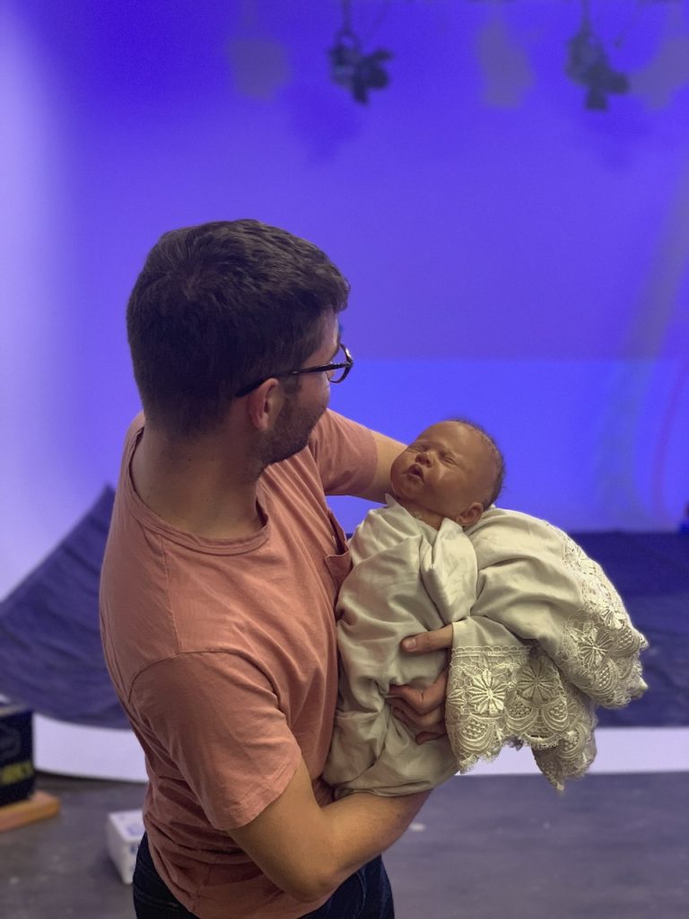 Man in glasses and peach shirt holding infant in white christening gown on purple-lit studio set