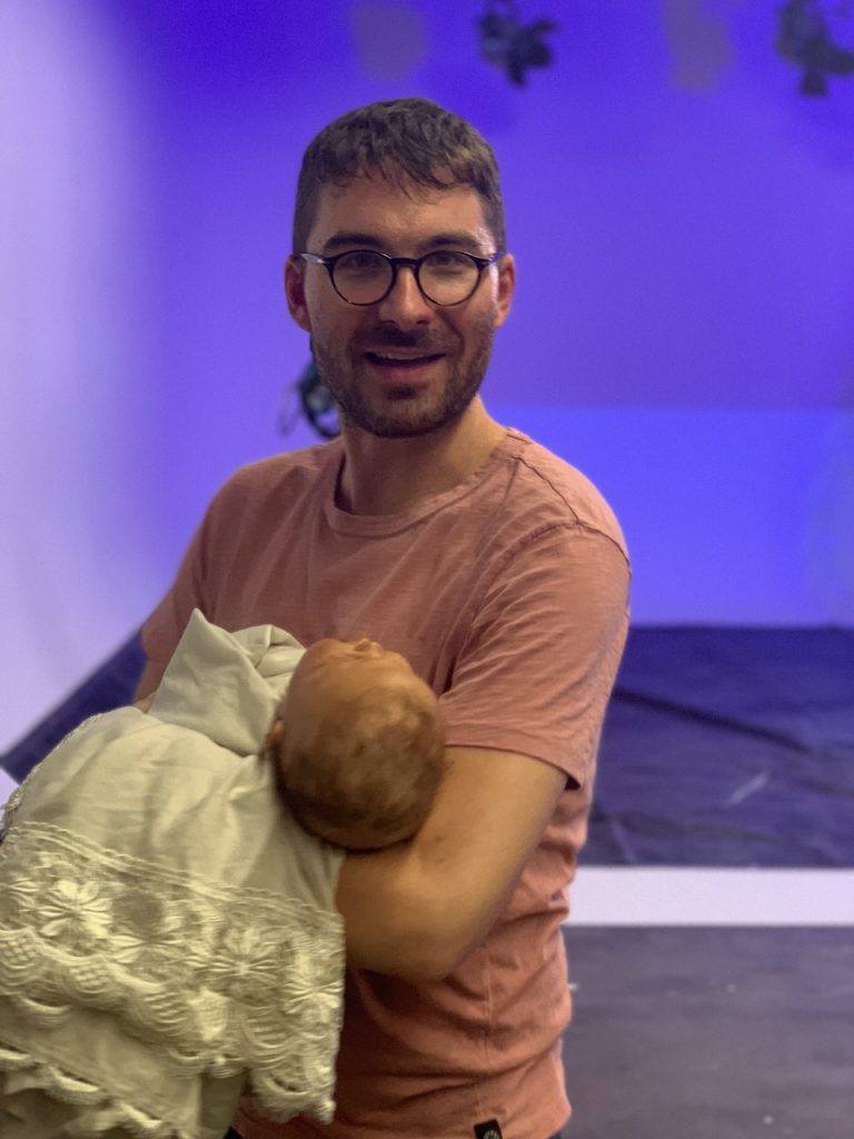 Director in glasses and tan shirt holding sleeping baby wrapped in yellow blanket on Tulsa Opera production set