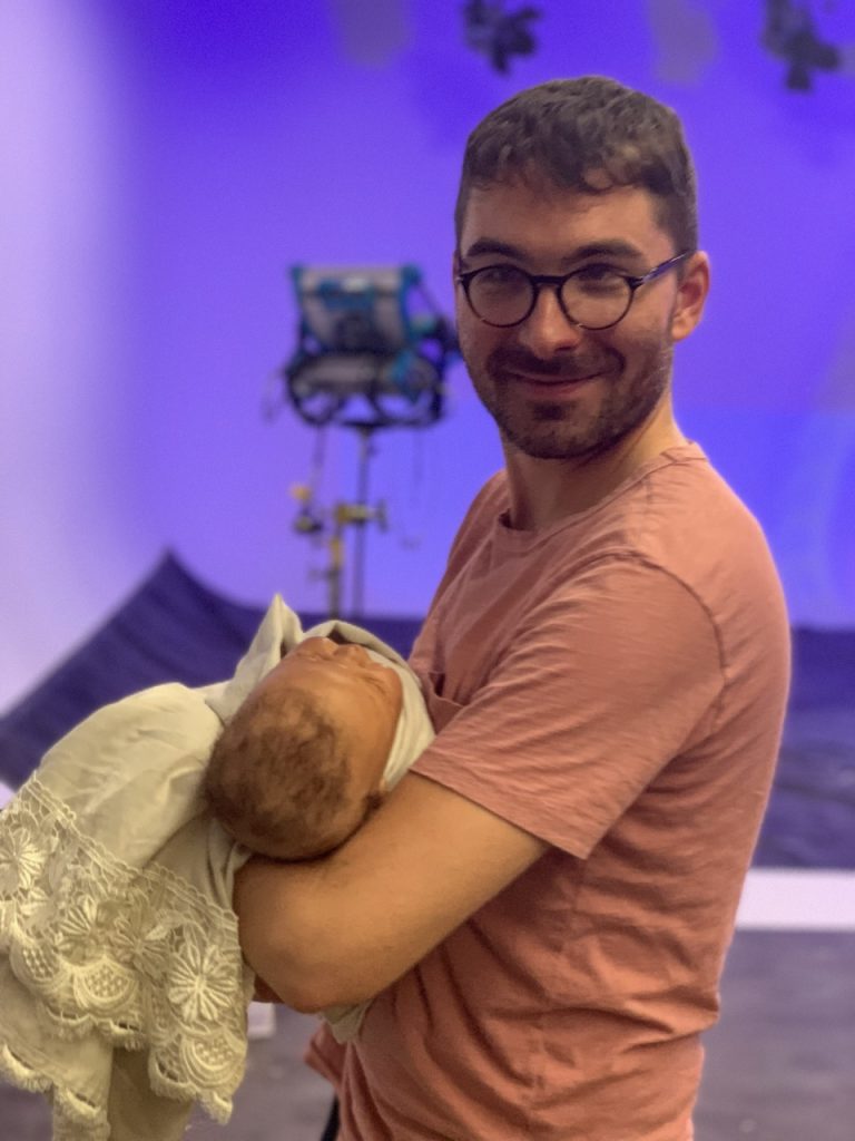 Man in peach shirt and glasses holding swaddled infant prop on film set with purple lighting, Tulsa Opera campaign