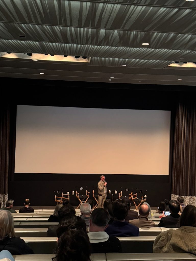 Director addresses audience from stage at theater screening, projection screen and seated attendees visible with James Kic...