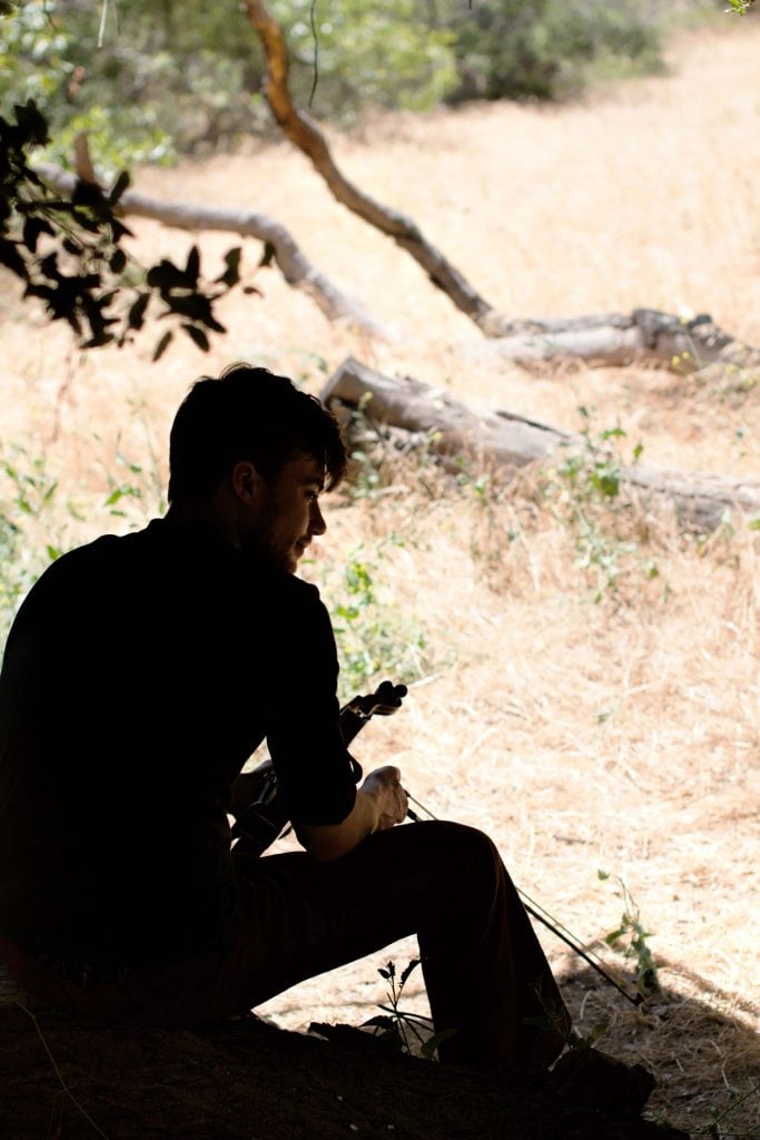 Silhouetted musician sits in shadow holding instrument outdoors, dry grass and tree branches in sunlit background