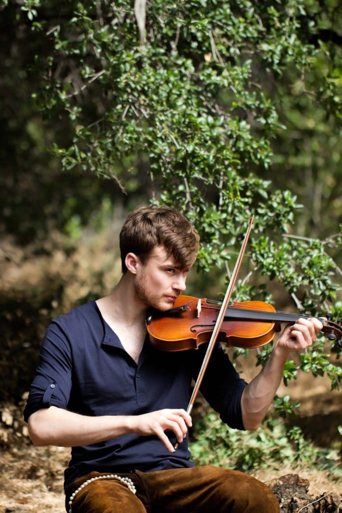 Violinist in black shirt playing violin outdoors among green foliage in natural forest setting