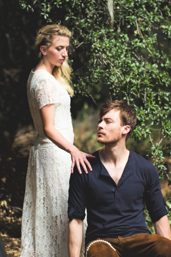 Woman in white lace dress standing beside man in dark shirt outdoors, surrounded by green foliage
