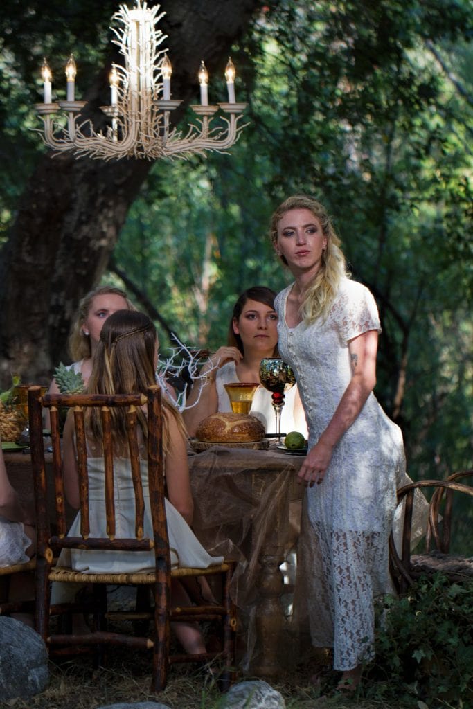Woman in white lace dress stands at rustic outdoor table set with bread and goblets, chandelier overhead, forest backdrop