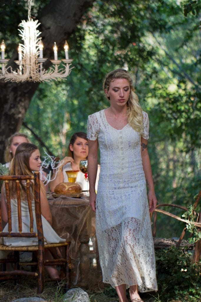 Blonde woman in white lace dress standing beside outdoor dining table under chandelier in forest, Branches music video