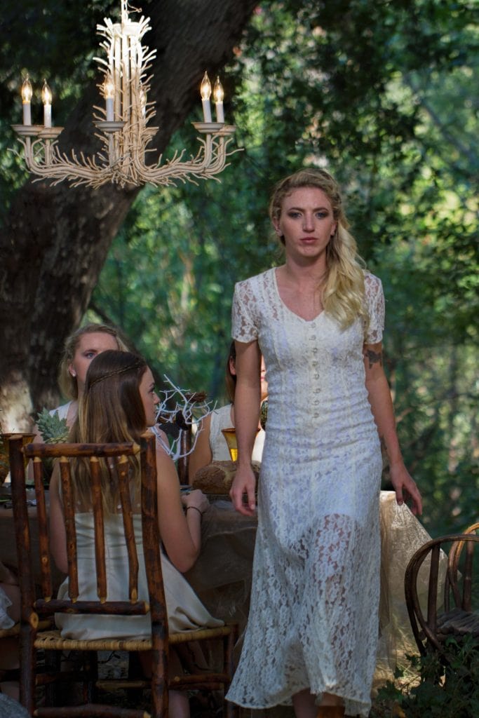 Woman in white lace gown stands beside rustic table with seated guests under ornate chandelier in forest setting