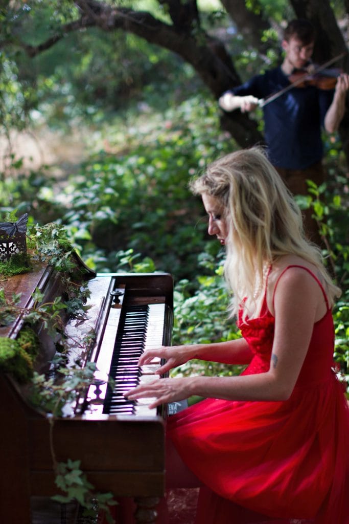 Blonde pianist in flowing red dress plays keyboard in forest, violinist performs in background. Branches music video.