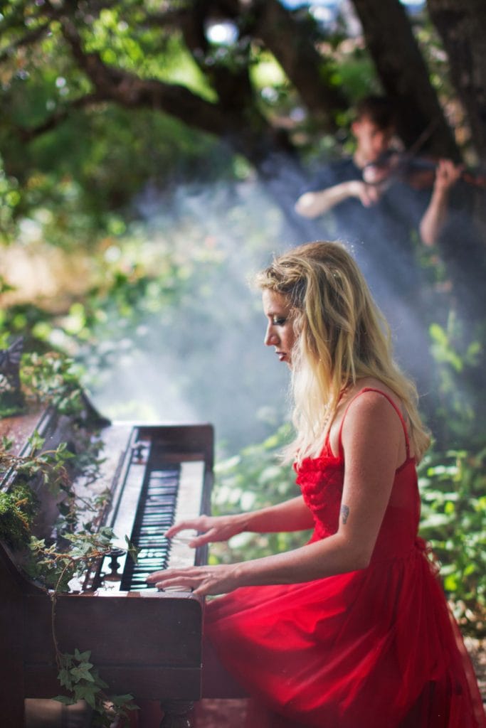 Blonde woman in red dress playing keyboard outdoors surrounded by greenery and smoke, Branches music video production