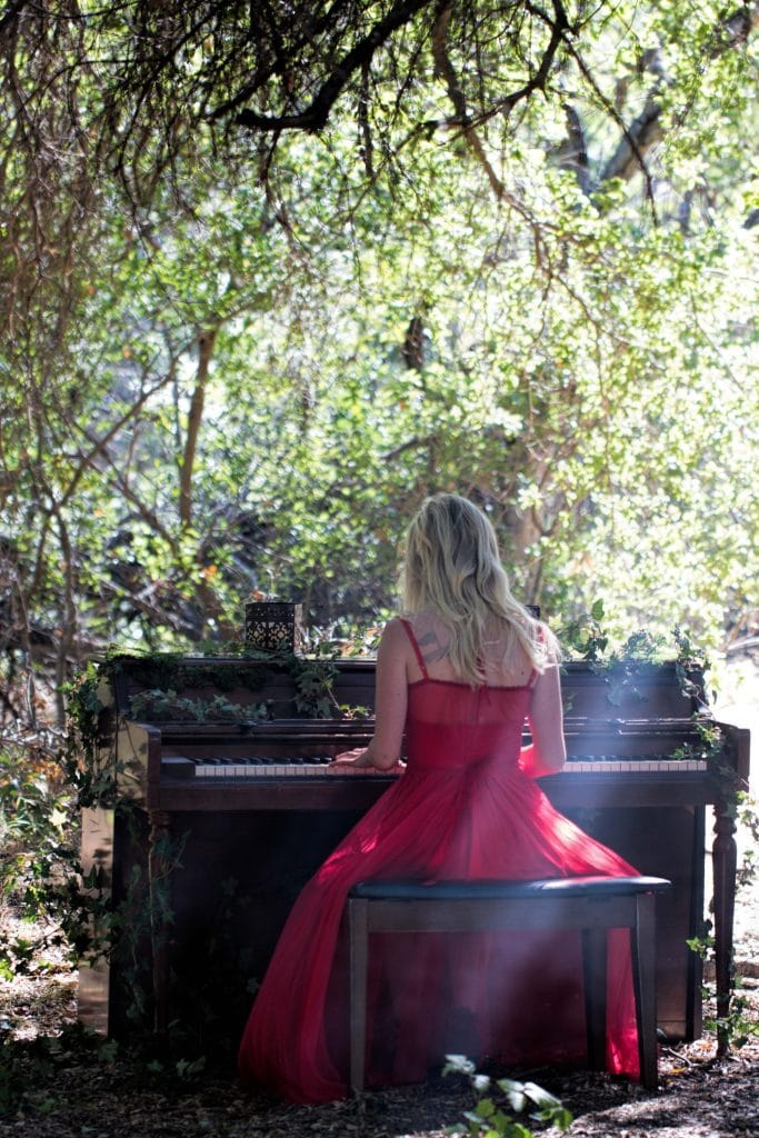 Blonde woman in flowing red gown seated at upright piano outdoors under canopy of trees, Branches music video shoot