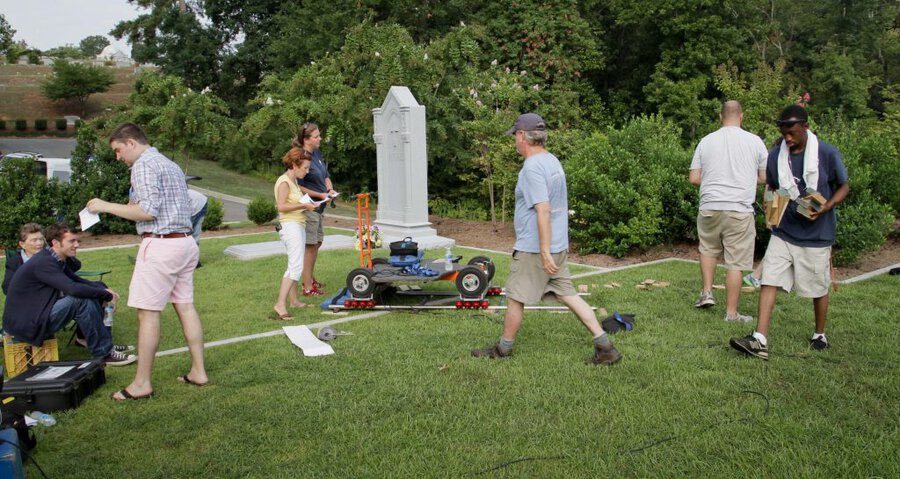 Film crew setting up wheeled dolly rig on grass near white monument, with crew members holding scripts and equipment
