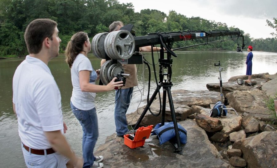 Crew operating camera jib on tripod by river, actor in blue coveralls on rocks, trees in background