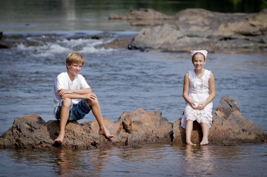 Two children in white clothing sitting on rocky outcrop in shallow river during film production