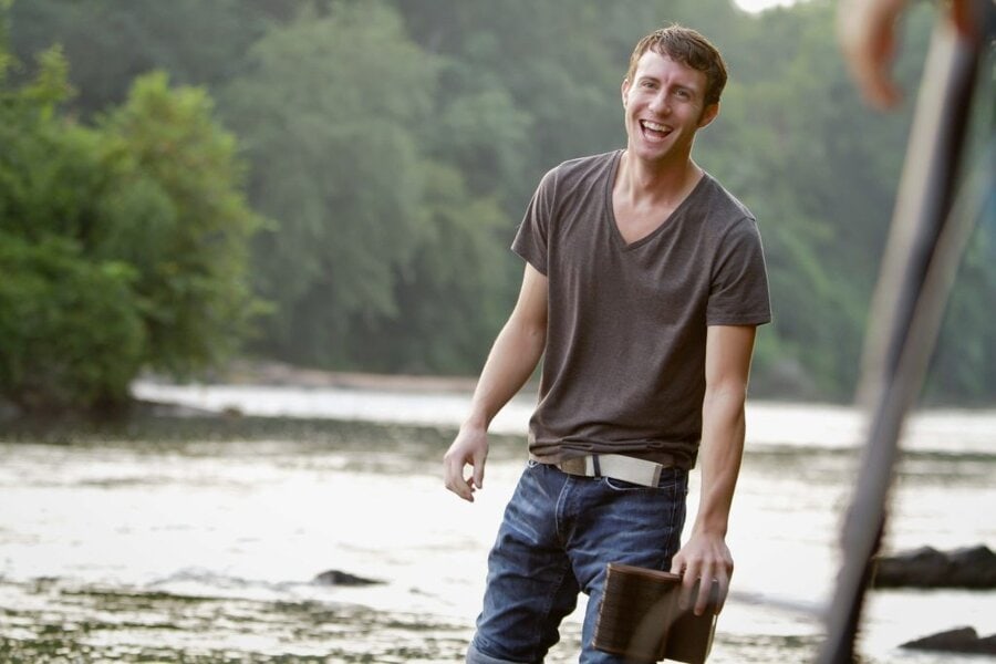 Young actor in brown t-shirt walks along river shore holding book, smiling during Final Acts film shoot in Macon